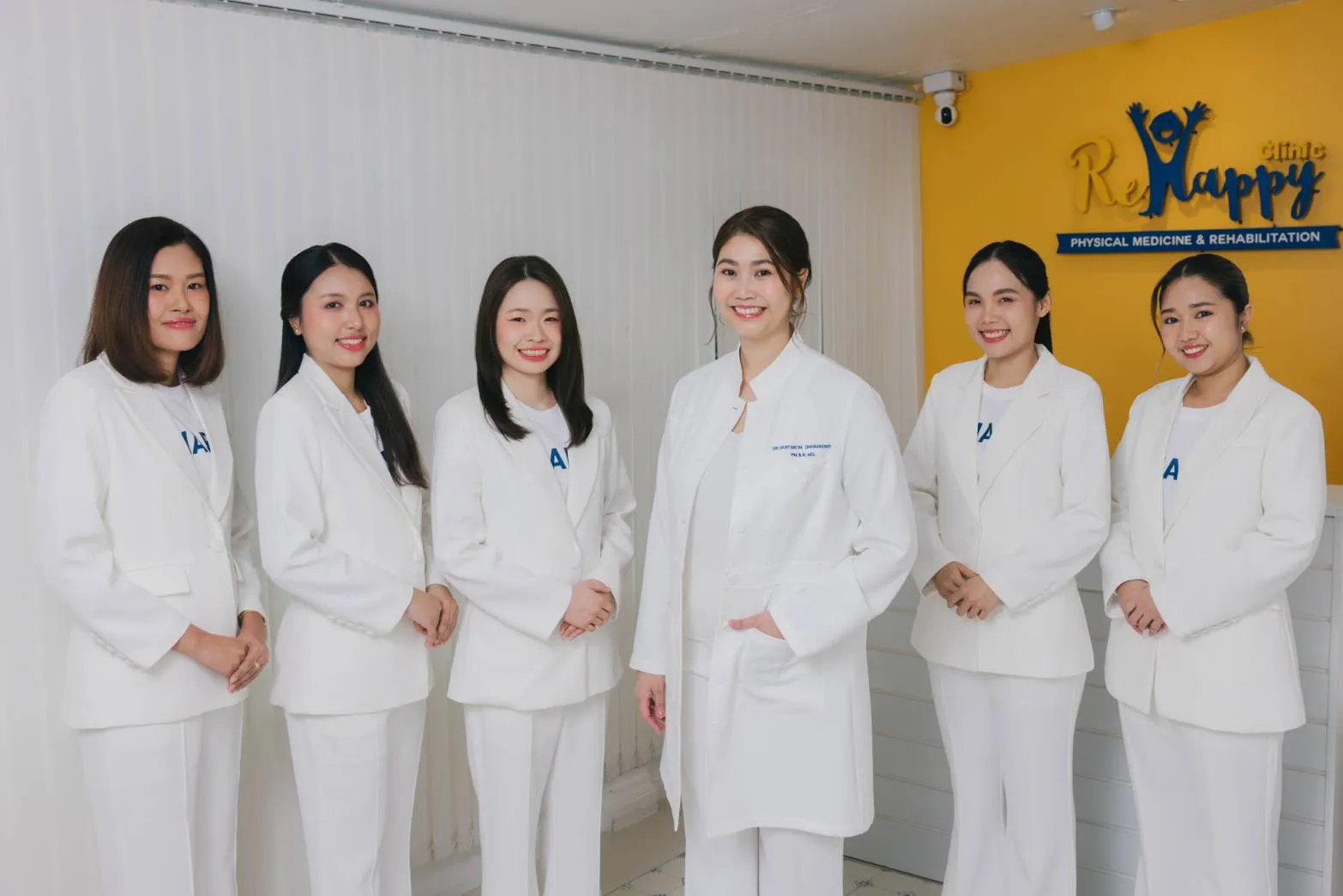ทีมงาน ReHappy Clinic ในชุดสูทสีขาว ยืนเรียงกันหน้าผนังโลโก้คลินิก